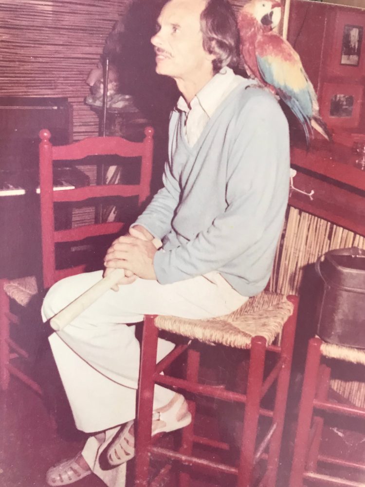 Ben's grandfather Dennis Boas sitting on a red stool with a macaw on his shoulder, piano behind him, at his restaurant Scandals
