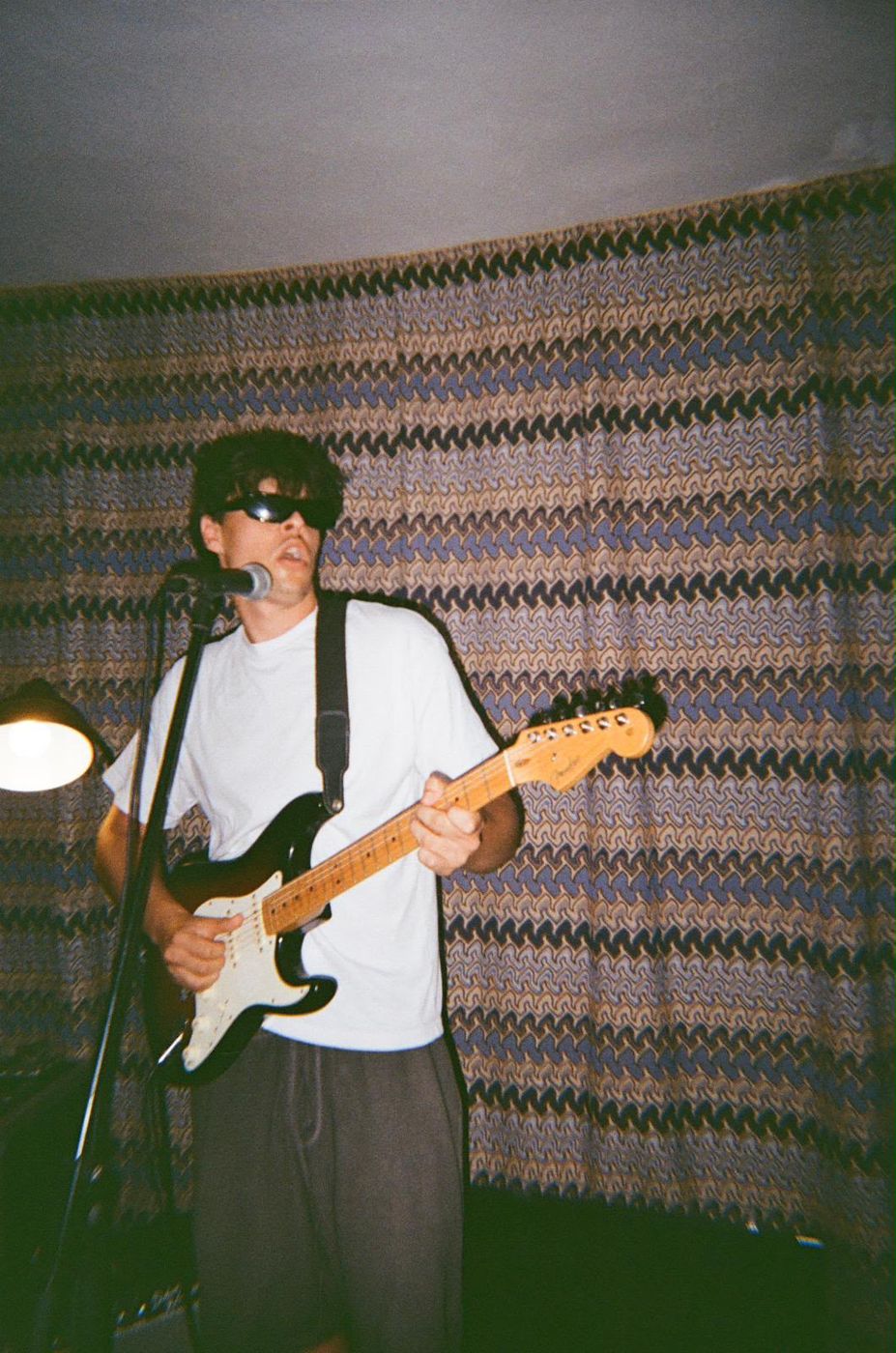 Ben Boas in sunglasses, singing and playing a black Stratocaster in front of a patterned curtain