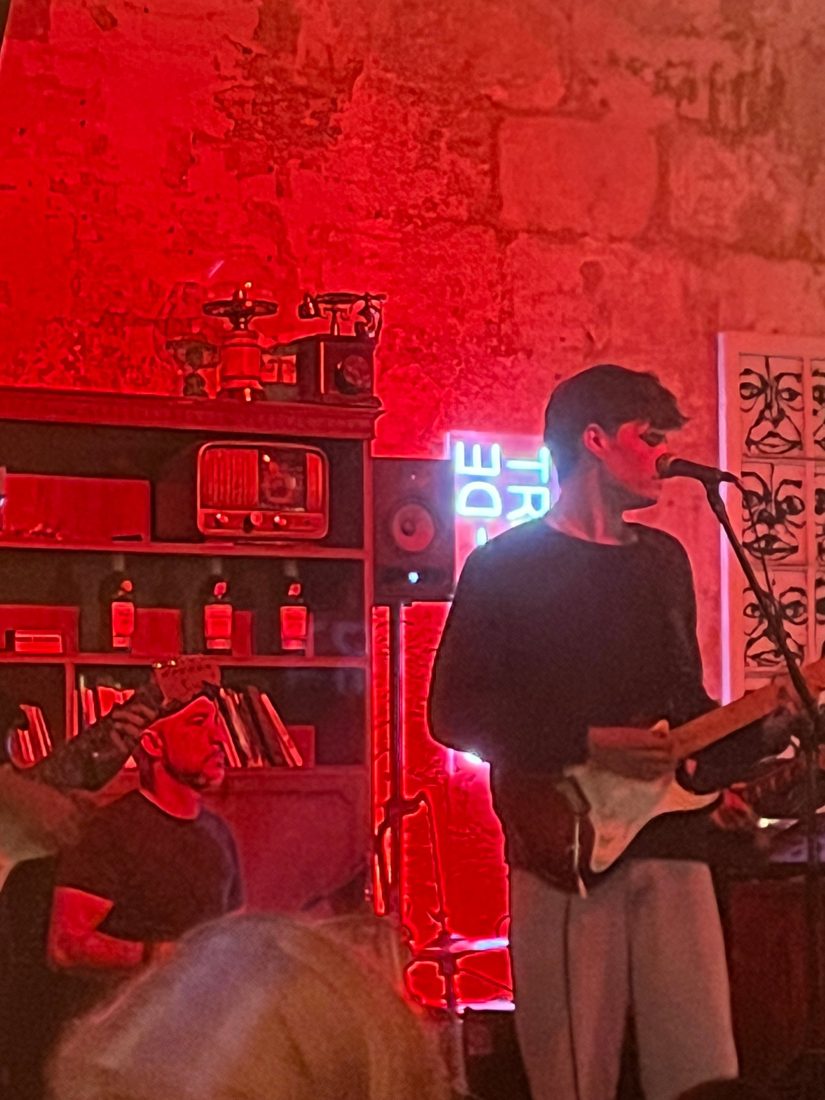 Ben singing and playing guitar under red light against an old stone wall at a jam session in Menorca